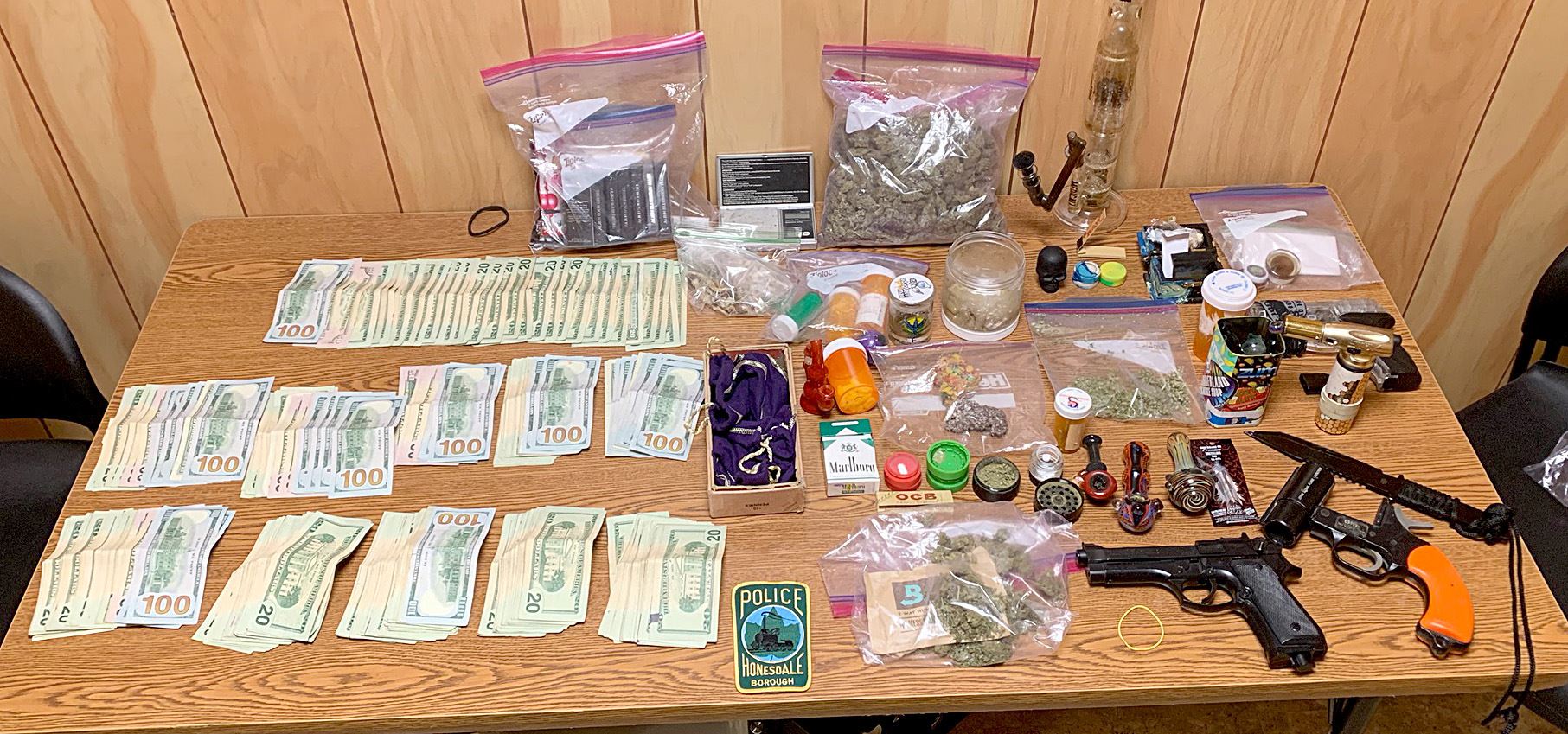 An array of US currency, marijuana and other drugs, along with paraphernalia and weapons on a table.